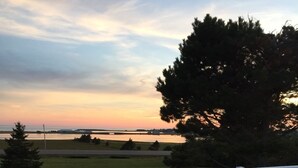 Eine Holzterrasse mit weißer Geländer, die auf eine Wiese mit Bäumen und eine entfernte Wassersicht im Sonnenuntergang blickt.