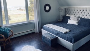 Ein Schlafzimmer mit großem Fenster, einem Bett mit grauem Polsterkopfteil und einer Holzbank.
