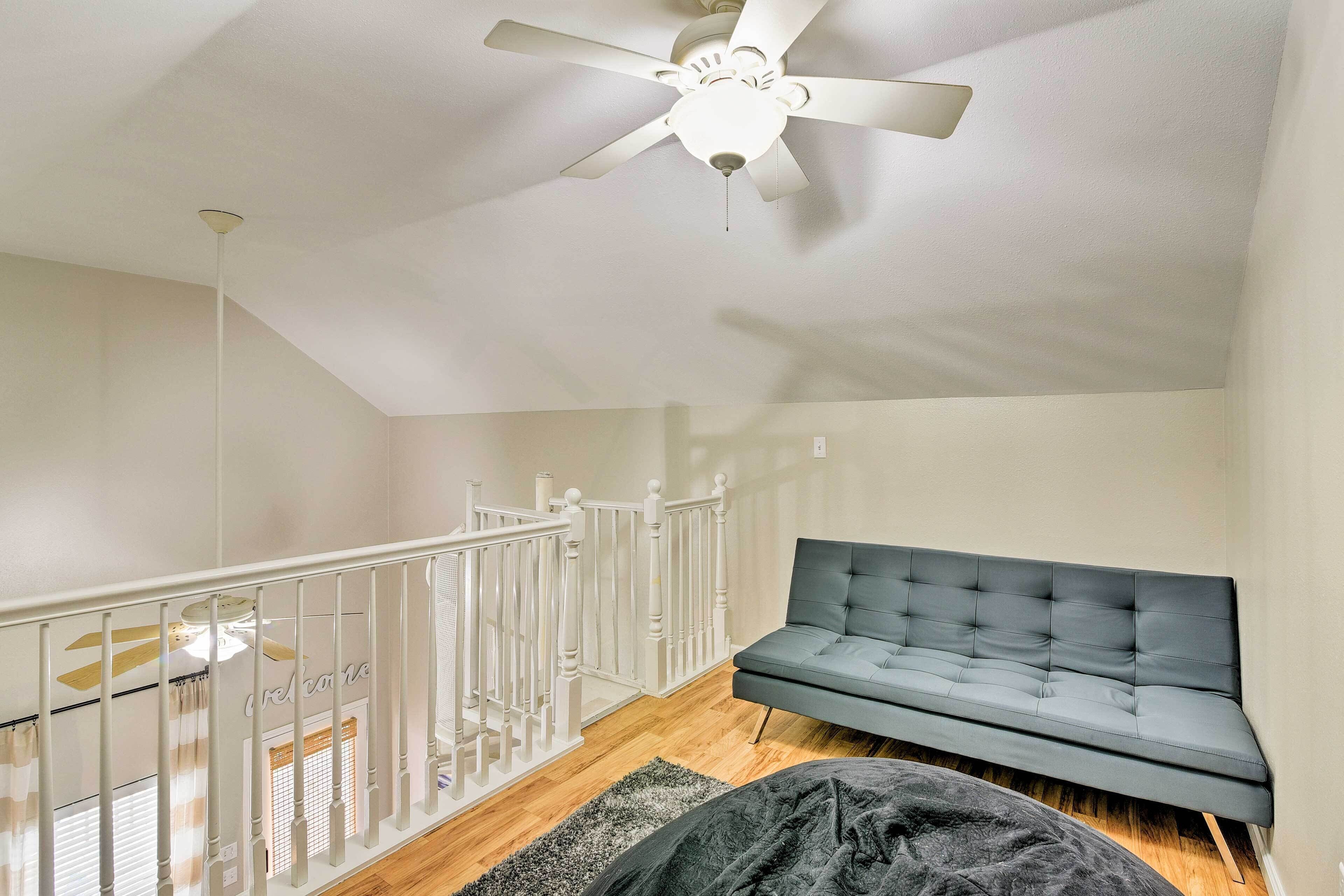 A modern loft bedroom with a grey sofa, a ceiling fan, and a white railing.