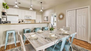 A dining area with a wooden table set for six, light blue chairs, and a kitchen in the background.