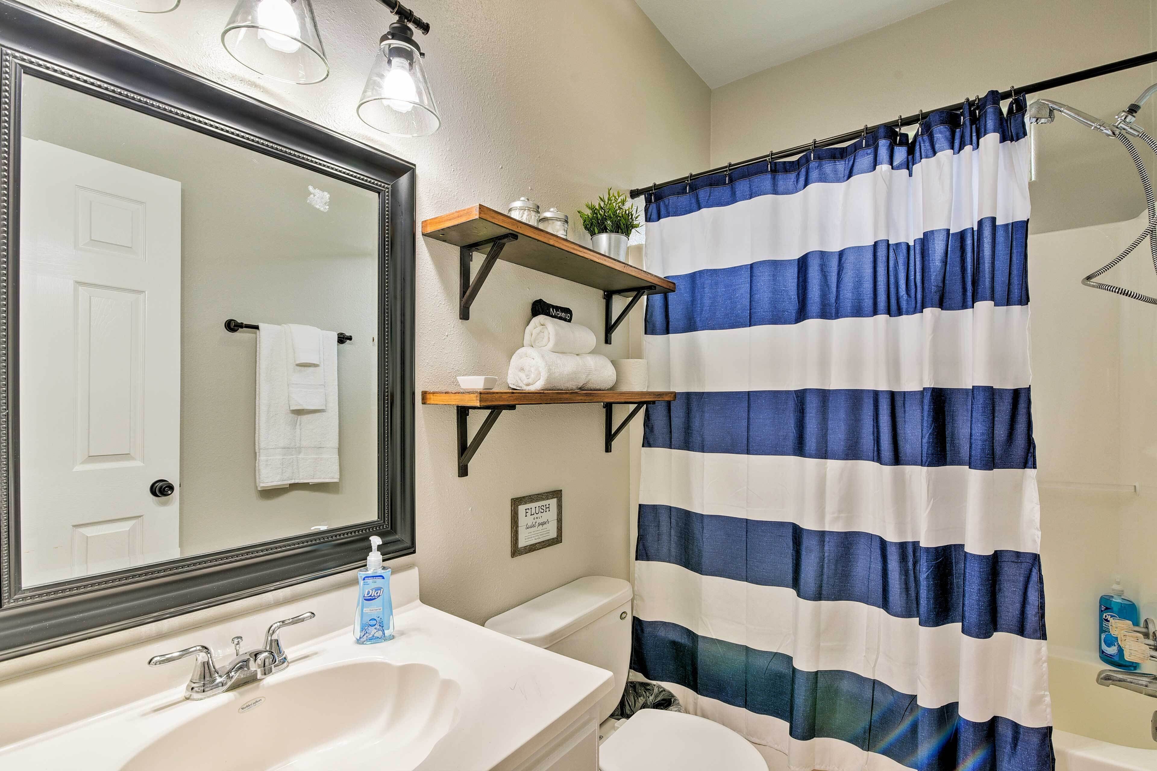 A bathroom with a white sink, a mirror, a toilet, a shower curtain, and a shelf with toiletries.