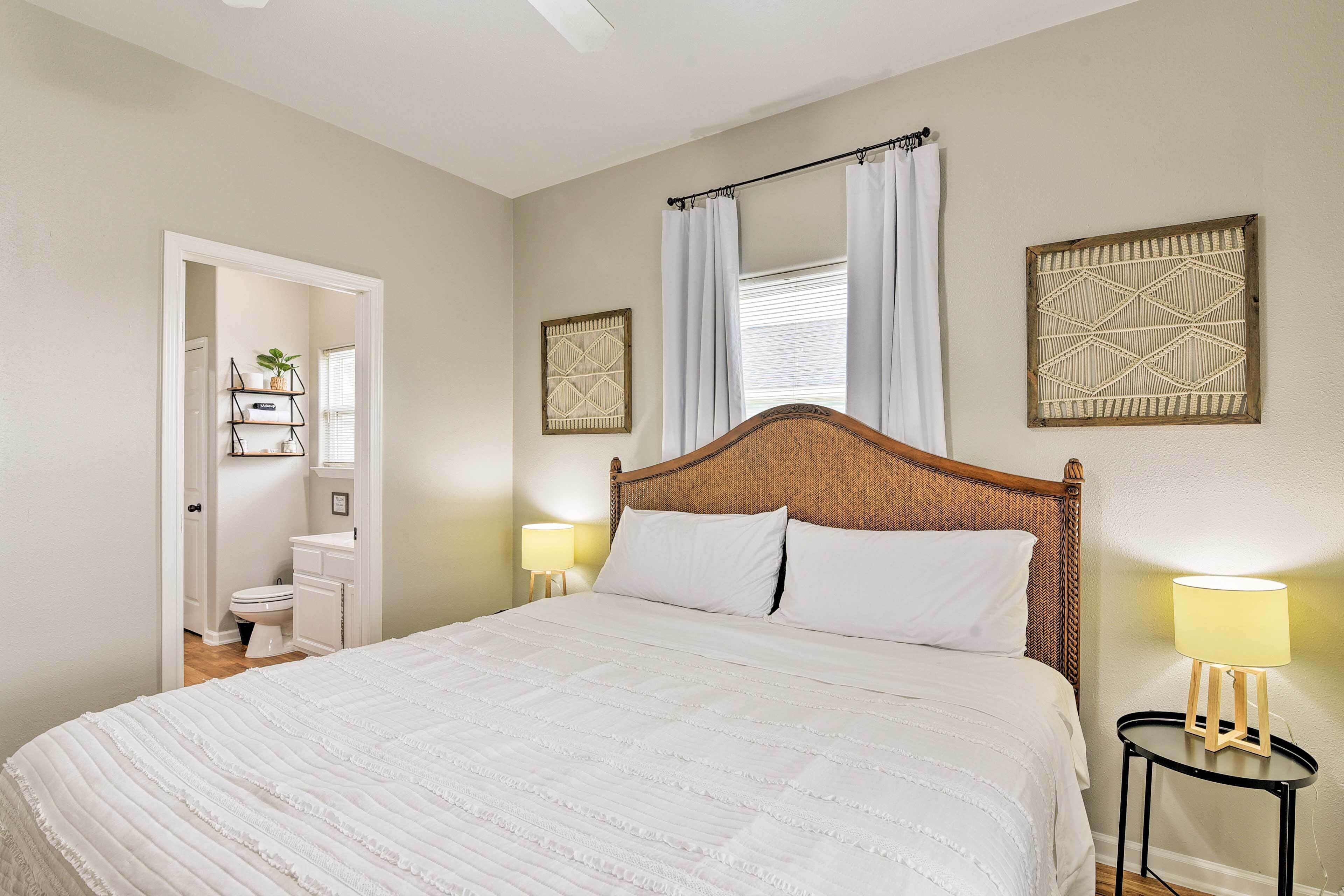 Quarto com cama grande, mesa de cabeceira com abajur, janela com cortinas e uma obra de arte pendurada na parede.