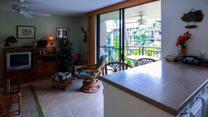 A living room with a TV, bookshelf, and a chair. There is a balcony with a view of greenery. - Oceanview Kailua-kona Condo With Private Balcony! (Kailua-Kona)