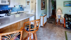A kitchen with white cabinets, a microwave, and a coffee maker. There are two wicker chairs with floral cushions. - Oceanview Kailua-kona Condo With Private Balcony! (Kailua-Kona)