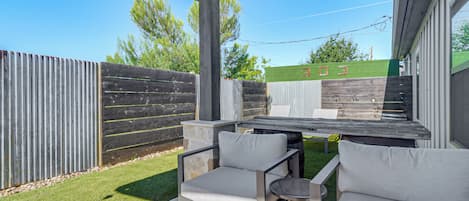 An outdoor patio with a covered seating area, a wooden table, and a view of a green lawn and a building in the background.