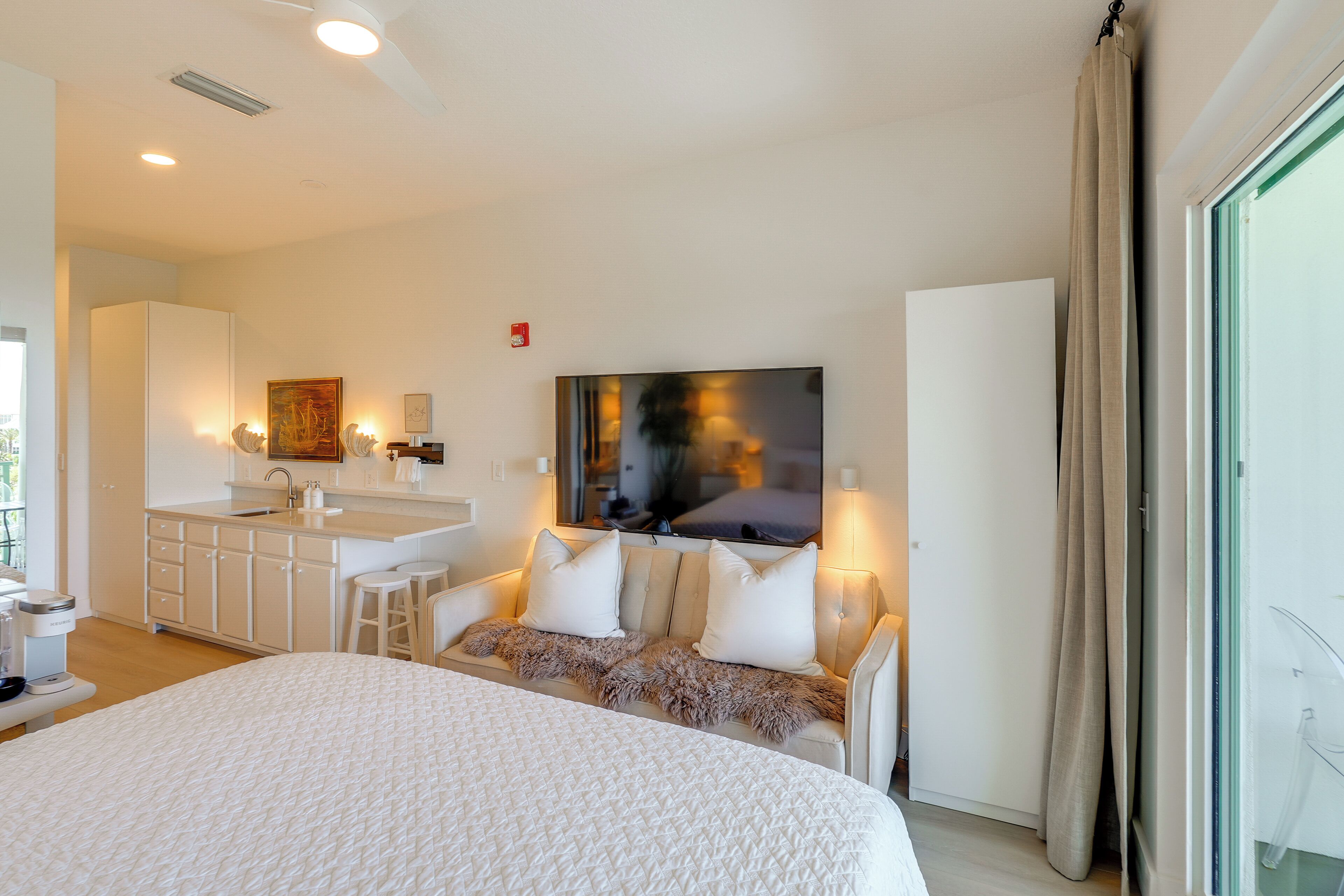 Una habitación de hotel moderna con una cama grande, un sofá, un televisor, una kitchenette y un ventanal amplio.