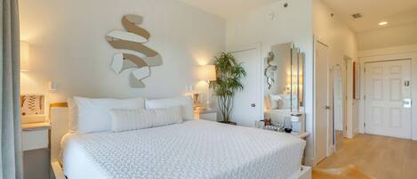 A bedroom with a large bed, bedside tables, a mirror, a potted plant, and a door leading to another room.