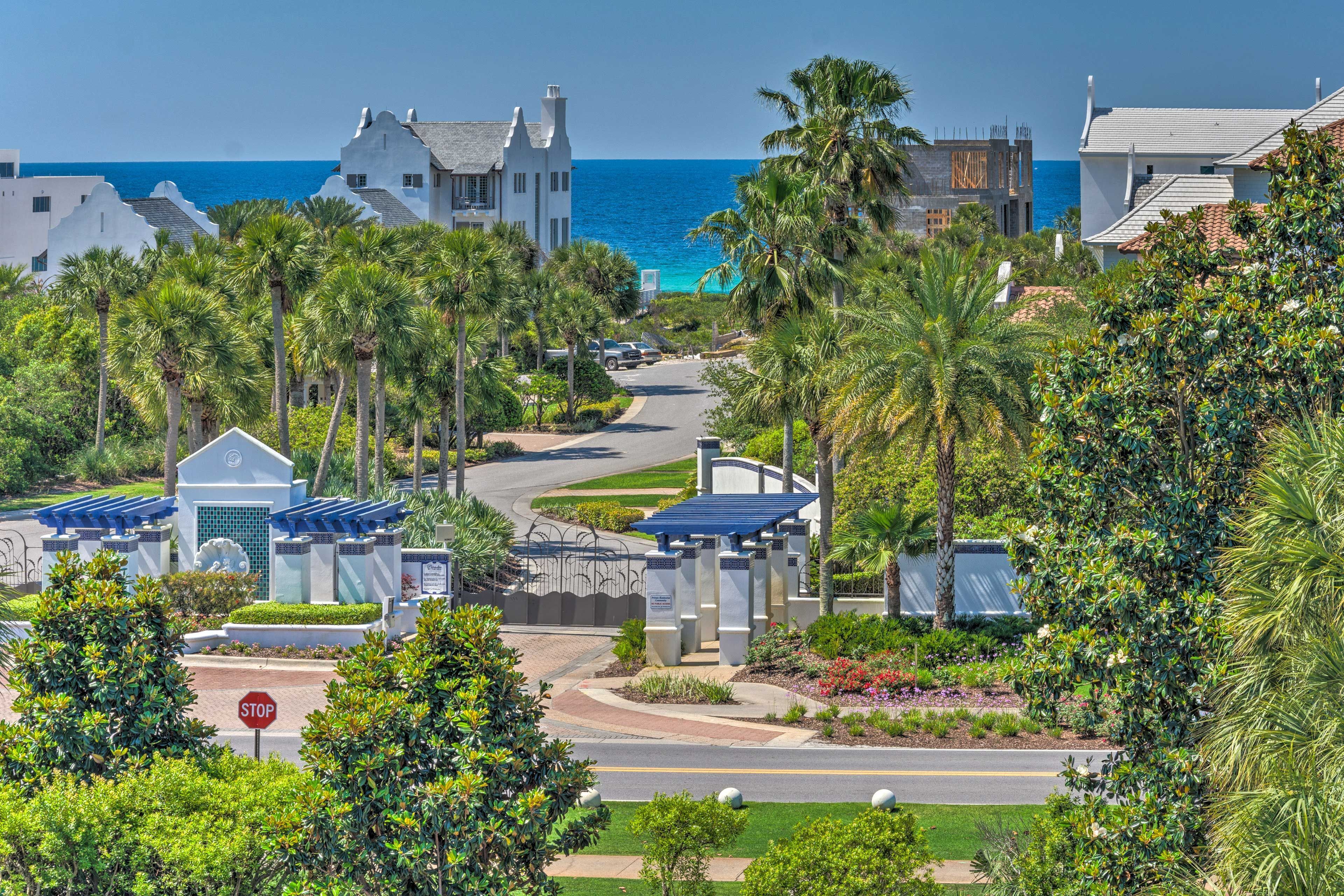 A coastal residential area with a road leading to a gated entrance, palm trees, and a view of the ocean.