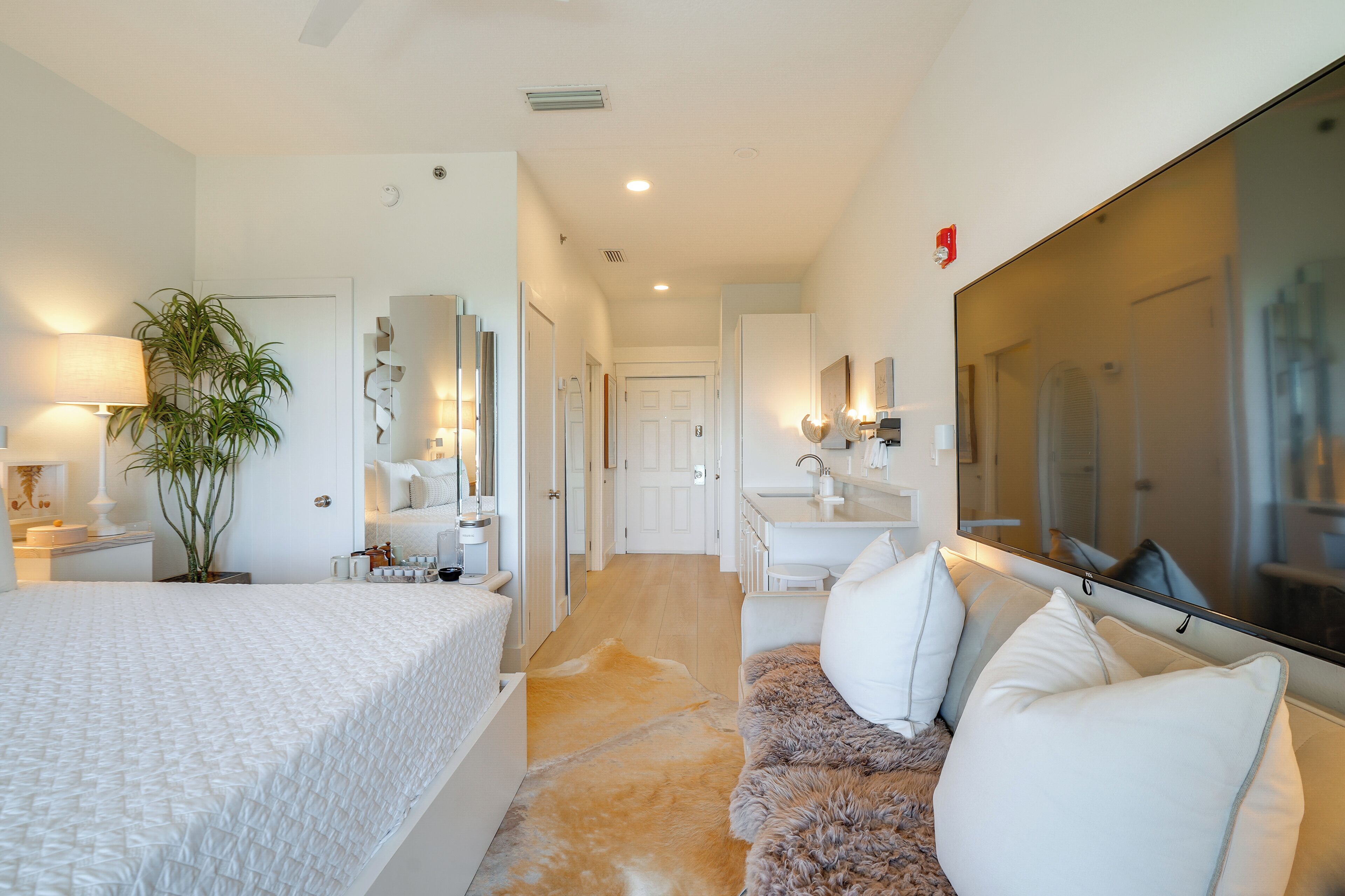 A modern bedroom with a large bed, a sitting area with a sofa, a mirror, a plant, and a bathroom with a sink and mirror.