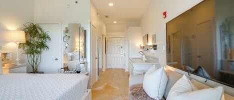 A modern bedroom with a large bed, a sitting area with a sofa, a mirror, a plant, and a bathroom with a sink and mirror.
