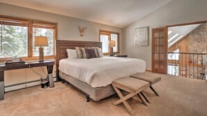 A bedroom with a large bed, a desk, and a bench. There are windows with curtains, a lamp, and a deer head decoration on the wall.