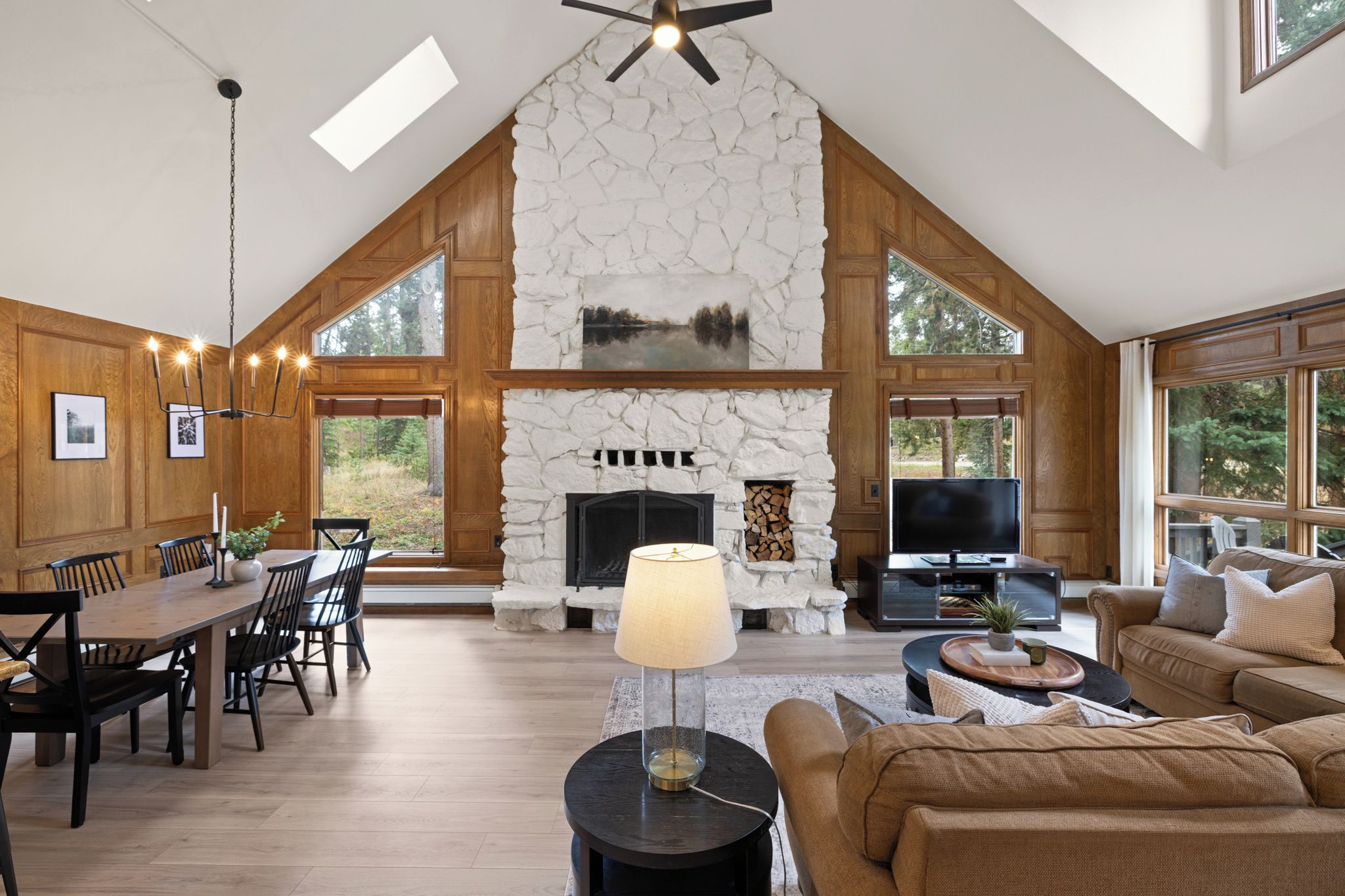 A spacious living area with a stone fireplace, wooden paneling, a dining set, and a large window offering a view of the outdoors.