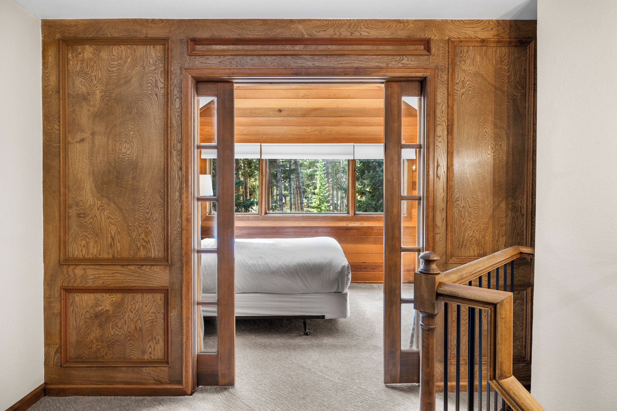 Quarto com cama, paredes de madeira e janela com vista para árvores.