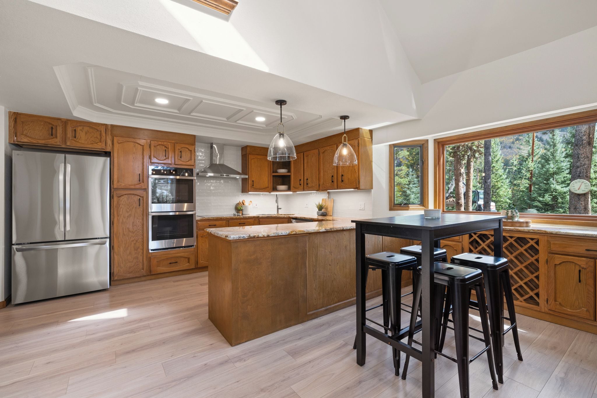 A modern kitchen with wooden cabinets, stainless steel appliances, a central island with stools, and large windows offering a view of trees.