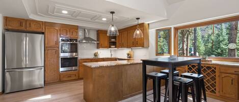 A modern kitchen with wooden cabinets, stainless steel appliances, a central island with stools, and large windows offering a view of trees.