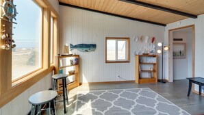 Une pièce avec des poutres apparentes en bois au plafond, de grandes fenêtres et une vue sur le paysage. On y trouve deux tabourets de bar, un tapis et des étagères ornées d’objets décoratifs.