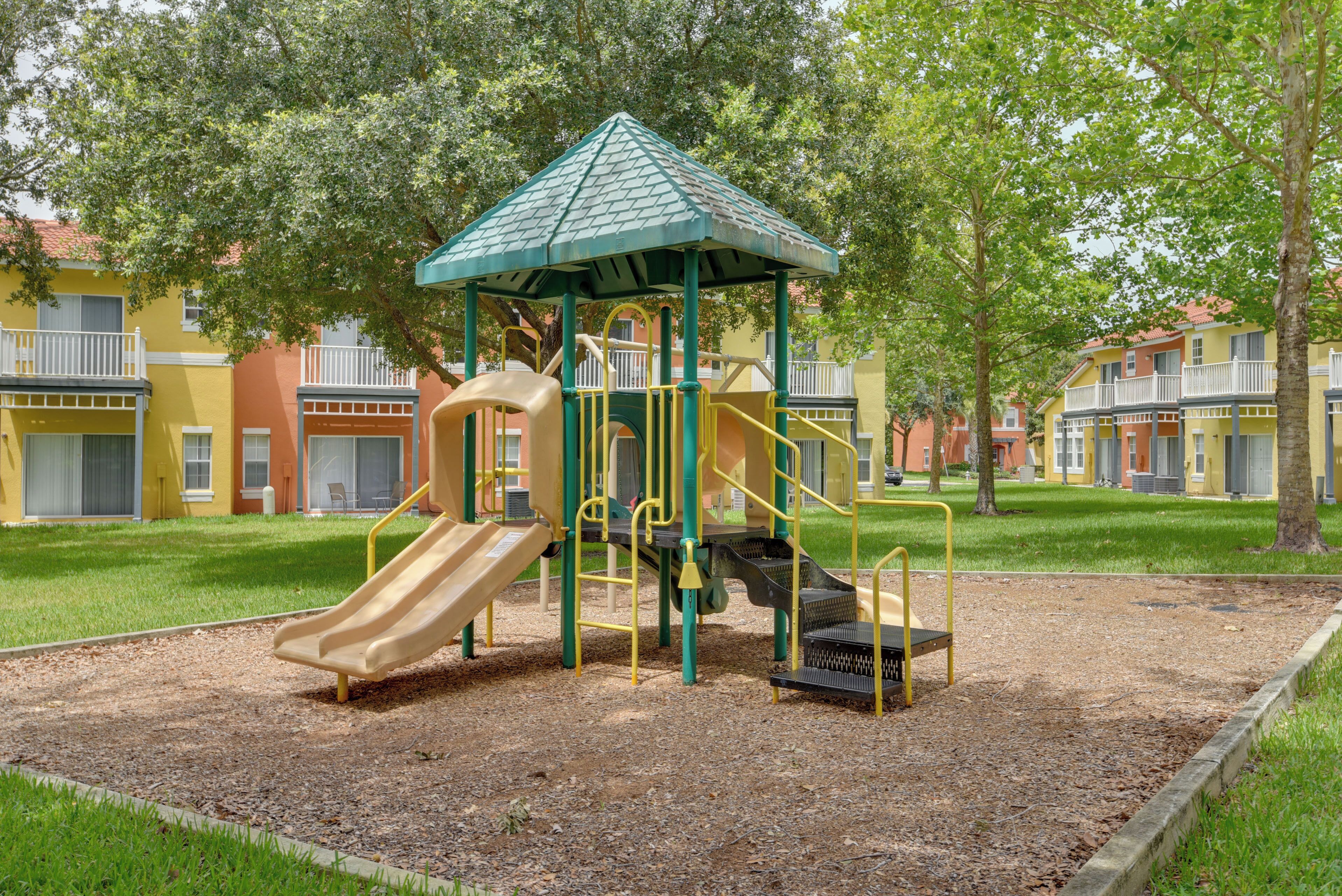 A playground with a slide and climbing structures, surrounded by a grassy area and trees.