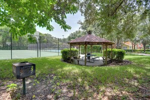 Um gazebo com uma mesa de piquenique e bancos, rodeado por grama e árvores.
