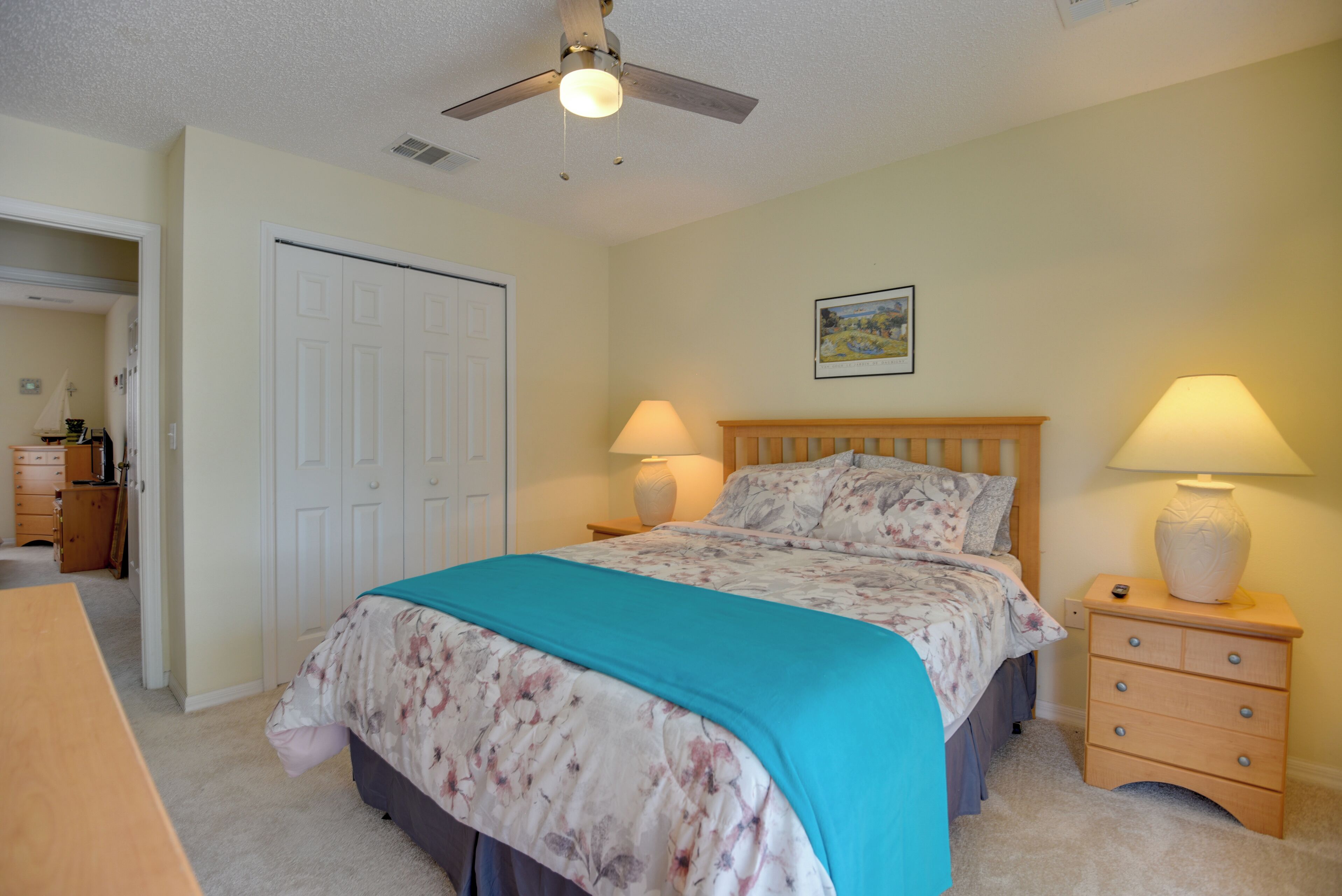 A bedroom with a bed, two bedside lamps, a ceiling fan, a wooden nightstand, and a door to another room.