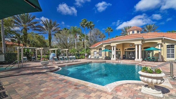 A swimming pool with lounge chairs and a covered seating area, surrounded by palm trees and a building with a red-tiled roof. - Near Disney & Epic Universe: Home w/ Resort Perks (Kissimmee)