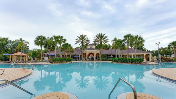 A large, clear swimming pool with a tiled deck, surrounded by palm trees and a building with a red roof in the background. - Private Cocktail Pool: Kissimmee Resort Retreat! (Kissimmee)