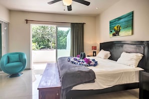 Un dormitorio moderno con una cama grande, un banco, una silla color turquesa y un cuadro de una escena tropical.