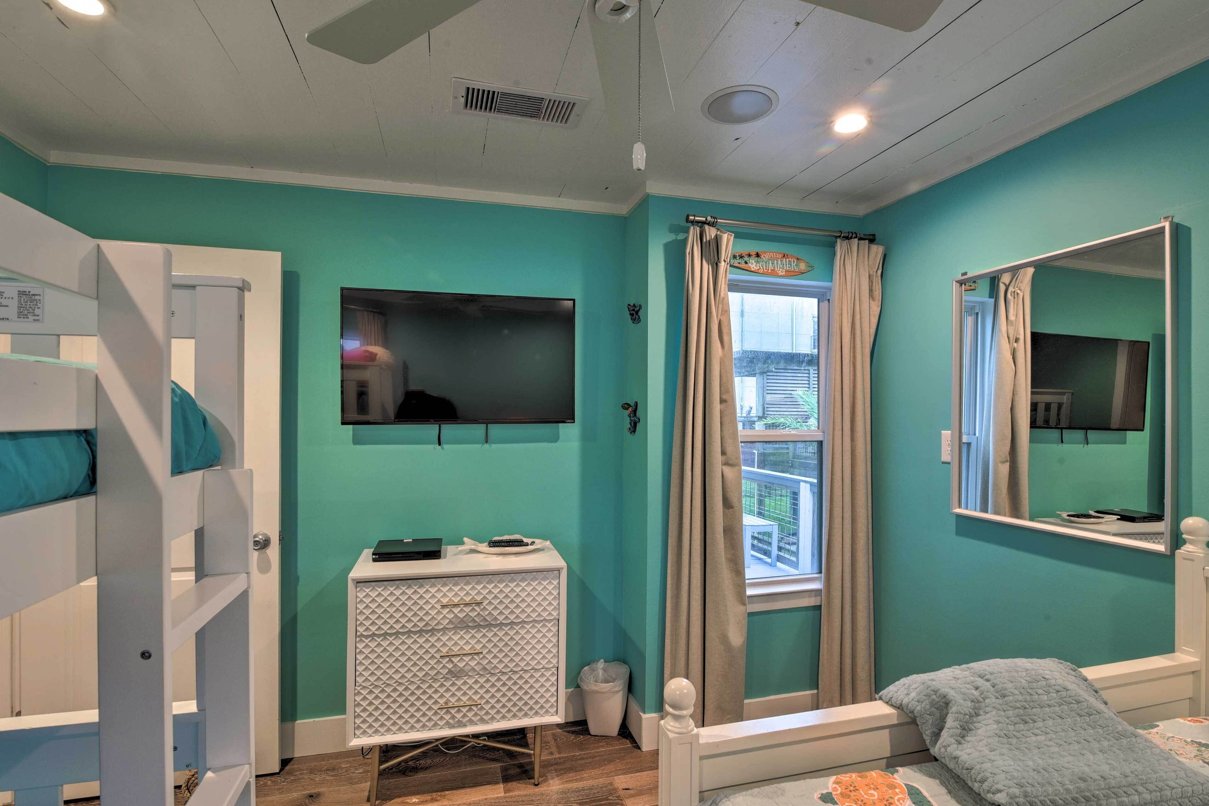 Quarto com paredes verde-água, uma cama beliche, uma cômoda pequena branca, uma janela com cortinas e um espelho.