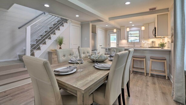 A modern dining area with a wooden table set for six, surrounded by cushioned chairs. A staircase with a metal railing is visible to the left. - Walk to Beach: Home in Galveston! (Galveston)