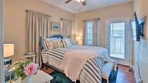A bedroom with a bed, curtains, a ceiling fan, a bedside table, a lamp, a flower vase, and a painting on the wall. - < 1 Mi to Stewart Beach: Gem in East End Galveston (Galveston)