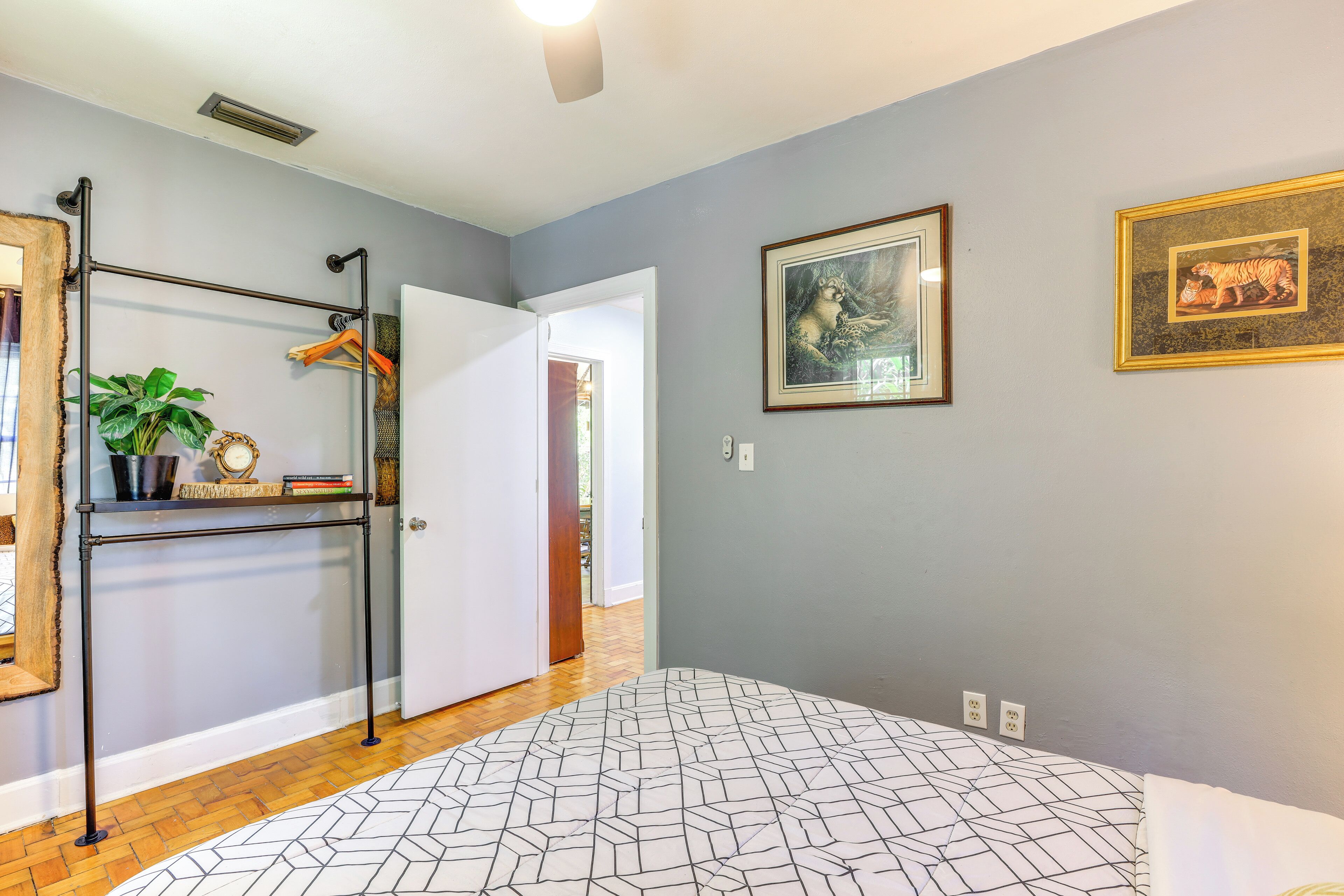 A bedroom with a patterned bedspread, a metal rack with clothes and a plant, and two framed pictures on the wall.