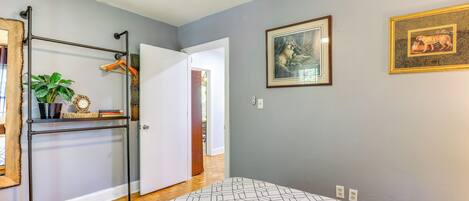 A bedroom with a patterned bedspread, a metal rack with clothes and a plant, and two framed pictures on the wall.