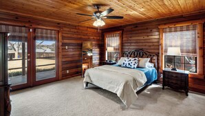 A wooden cabin bedroom with a bed, a nightstand, a ceiling fan, and large windows. - Lakefront Cabin + Guest House Near Granbury Square (Granbury)
