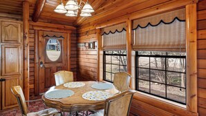 A wooden cabin interior with a dining table, chairs, and a large window with blinds. - Lakefront Cabin + Guest House Near Granbury Square (Granbury)