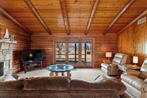 A log cabin interior with a stone fireplace, a television, leather sofas, and a glass coffee table.