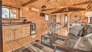 A spacious log cabin with a kitchen, dining area, and living room. It features wooden floors, a ceiling fan, and a large wheel on the wall.
