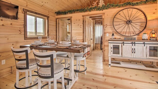 A wooden cabin interior with a dining table, chairs, a large wheel, and a kitchen area.