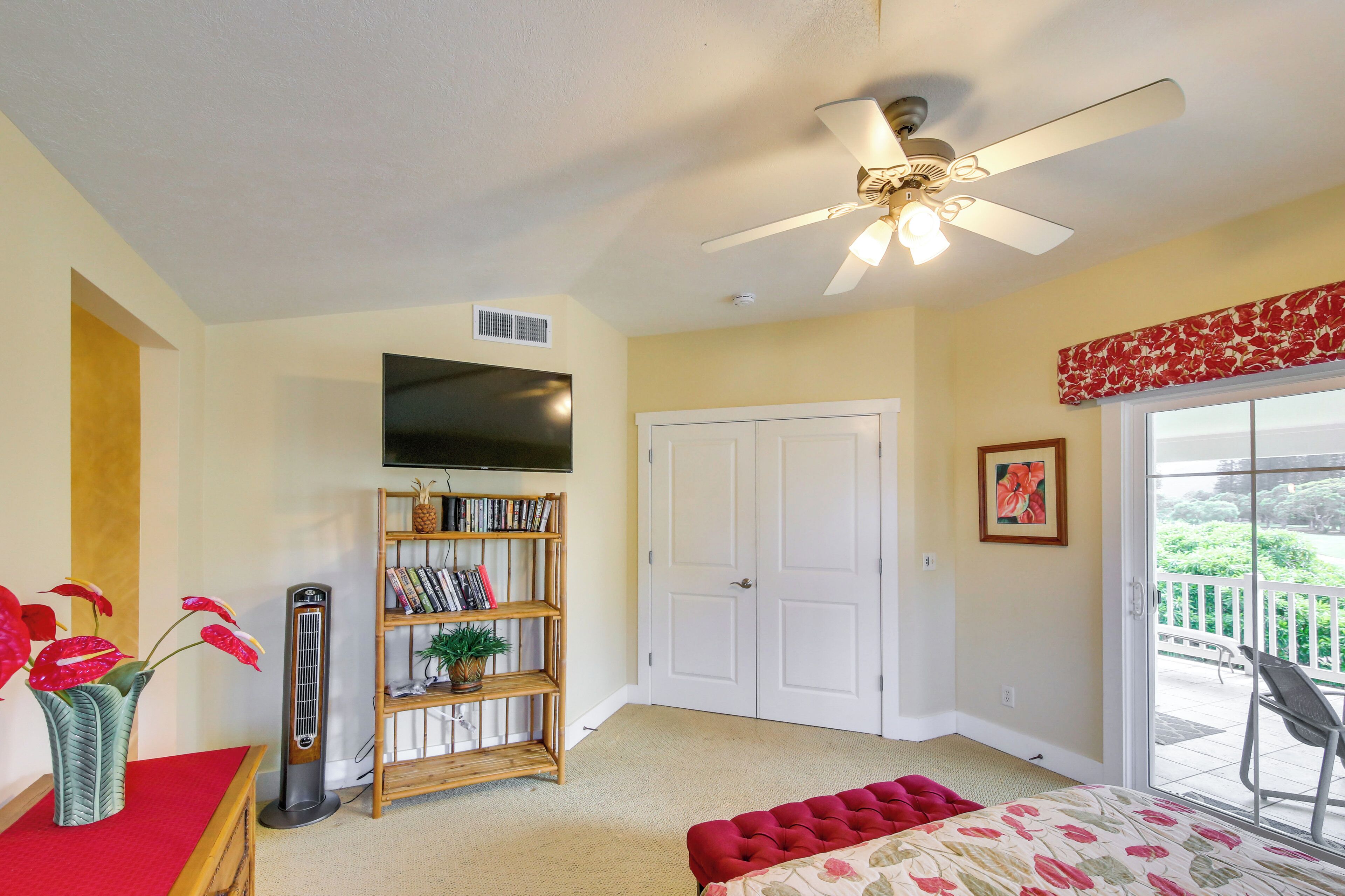 A bedroom with a ceiling fan, a bookshelf, a TV, a sliding glass door to a balcony, and a red ottoman.