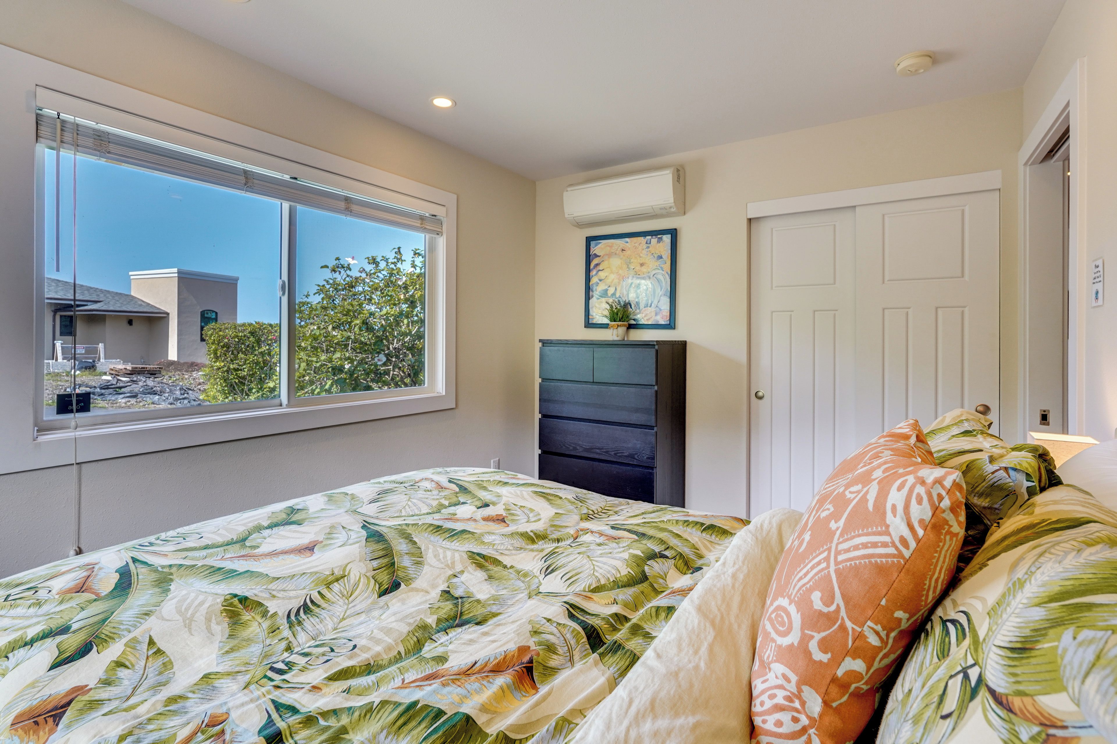 A bedroom with a large window, a bed with a tropical leaf pattern bedspread, a dresser, and a painting on the wall.