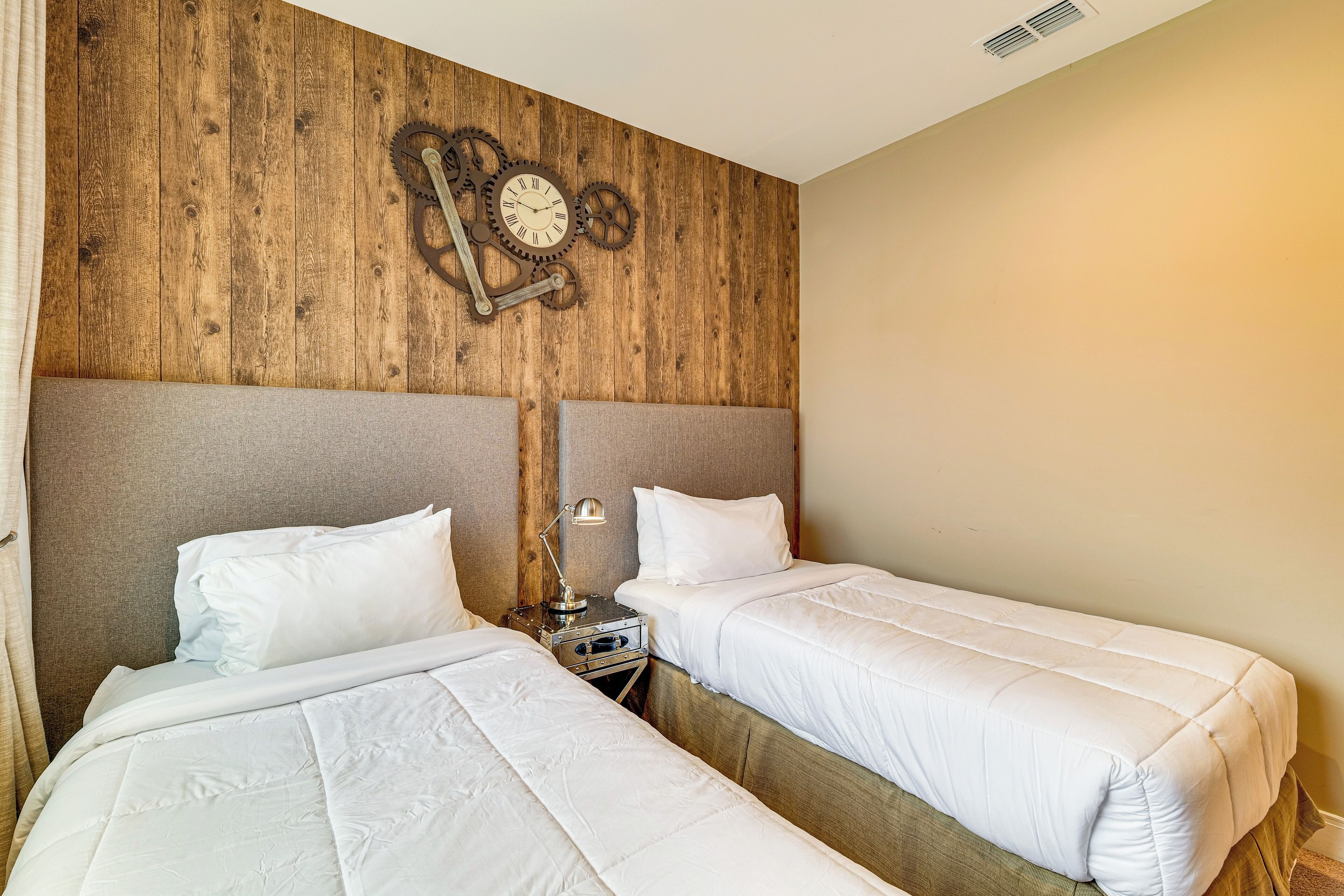 Una habitación con dos camas, paneles de madera en las paredes, un reloj y una mesita de noche con lámpara.