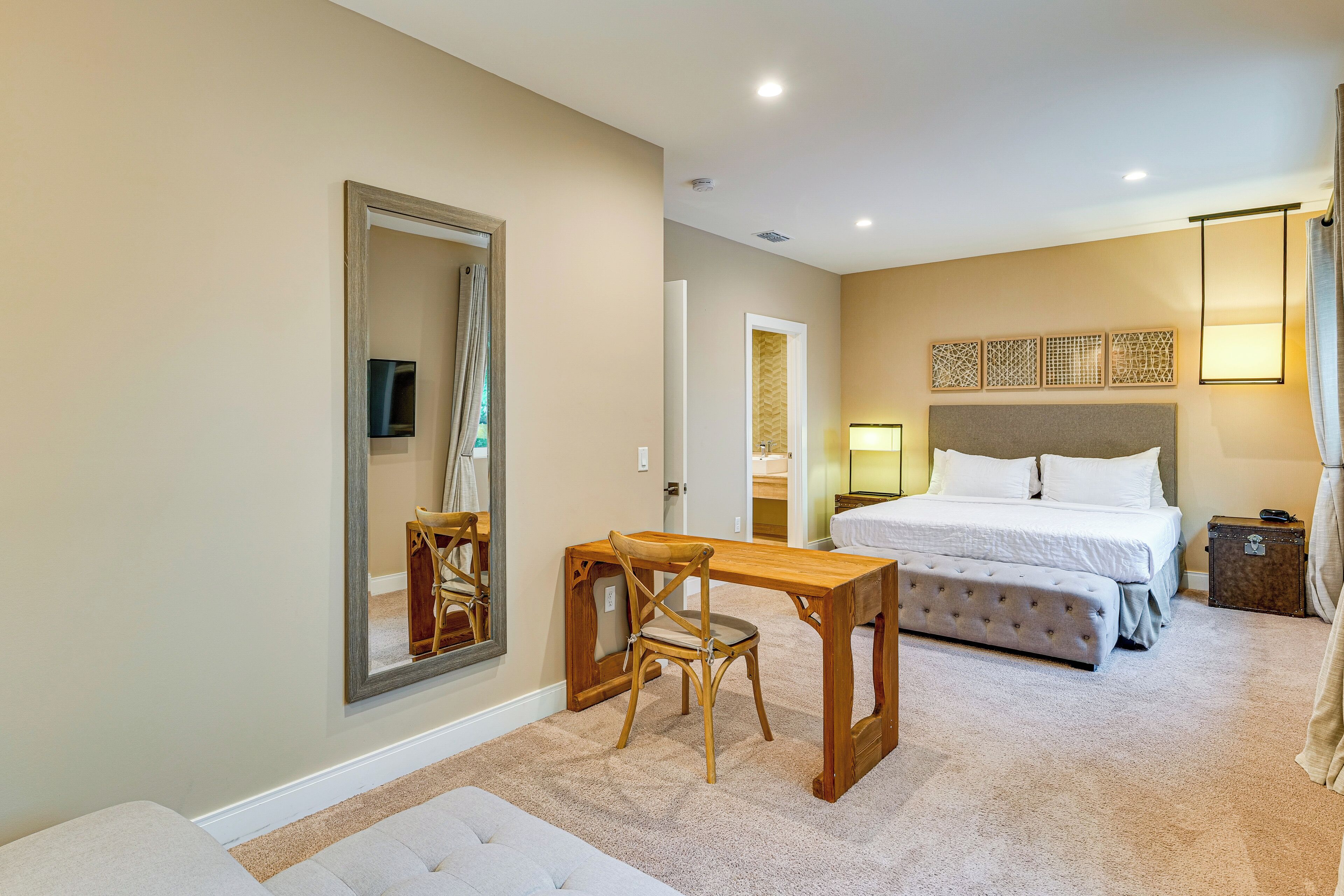 A hotel room with a bed, a wooden desk, a chair, a small table, and a mirror.