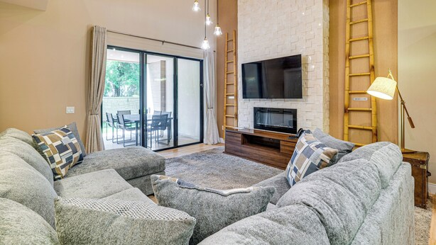 Ein modernes Wohnzimmer mit einem Ecksofa, einem Flachbildfernseher, einem Kamin und einem großen Fenster mit Blick ins Freie.