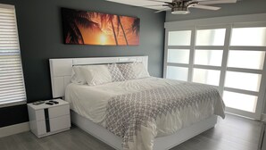Une chambre à coucher avec un grand lit, une table de chevet, un ventilateur de plafond et une fenêtre avec des stores.
