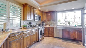 A kitchen with wooden cabinets, stainless steel appliances, and a large window offering a view of palm trees. - Palm Desert Escape w/ Patio & Shared Pool! (Palm Desert)