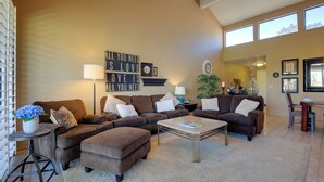 A spacious living room with a sectional sofa, ottoman, and coffee table. There is a wall-mounted sign that reads "All you need is love." The room has large windows, a fireplace, and a dining area with a table and chairs.
