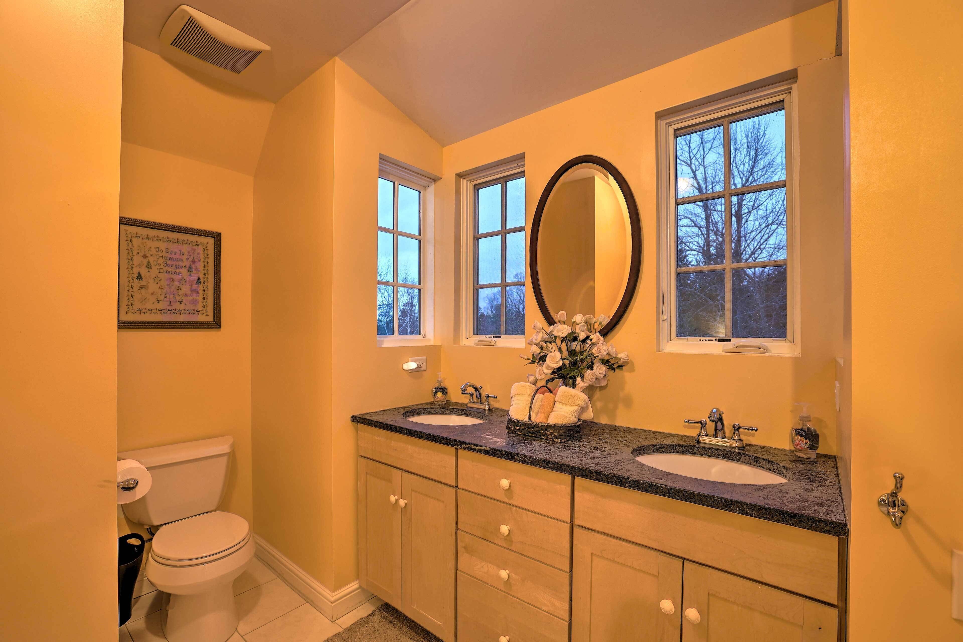 A bathroom with two sinks, a toilet, and a large mirror. There are windows above the sinks and a framed picture on the wall.