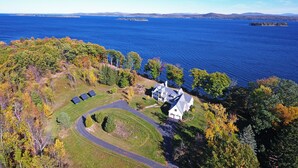 A large house with a driveway and solar panels, surrounded by trees and a lake. - Stunning South Hero Home on Lake Champlain w/ View (South Hero)