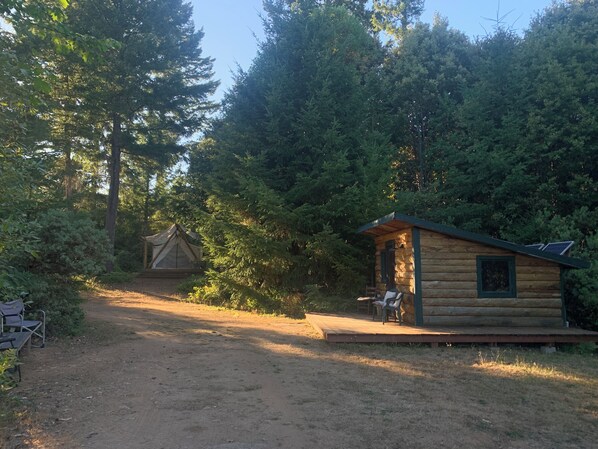 Una cabaña rústica con terraza de madera y una pequeña mesa, rodeada de un bosque frondoso.