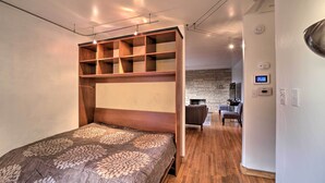 A modern bedroom with a wooden bed, built-in shelves, a brick wall, and a small seating area in the background. - Tulsa Rental Home - Walk to Philbrook Museum (Tulsa)