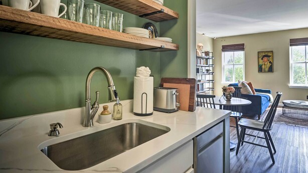 A kitchen with a large sink, wooden shelves with dishes, a toaster, and a dining area with a blue sofa and a table. - Classic Fells Point Condo on Broadway Square! (Baltimore)