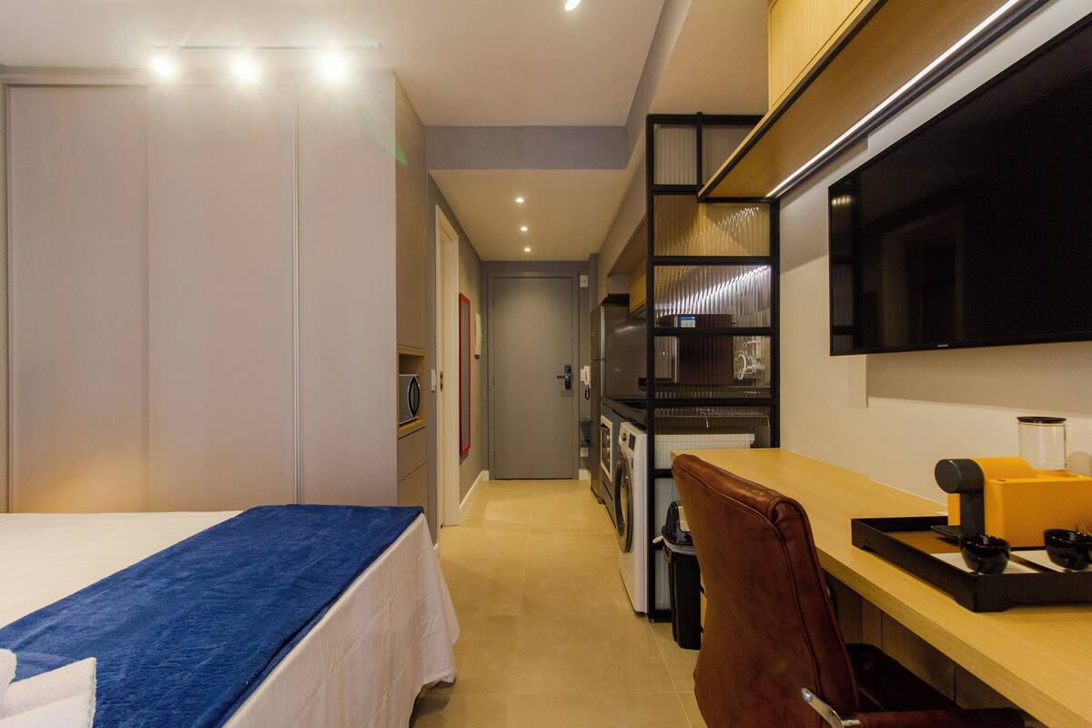 A hotel room with a bed, a desk, a chair, a TV, a washing machine, and a small kitchen area.